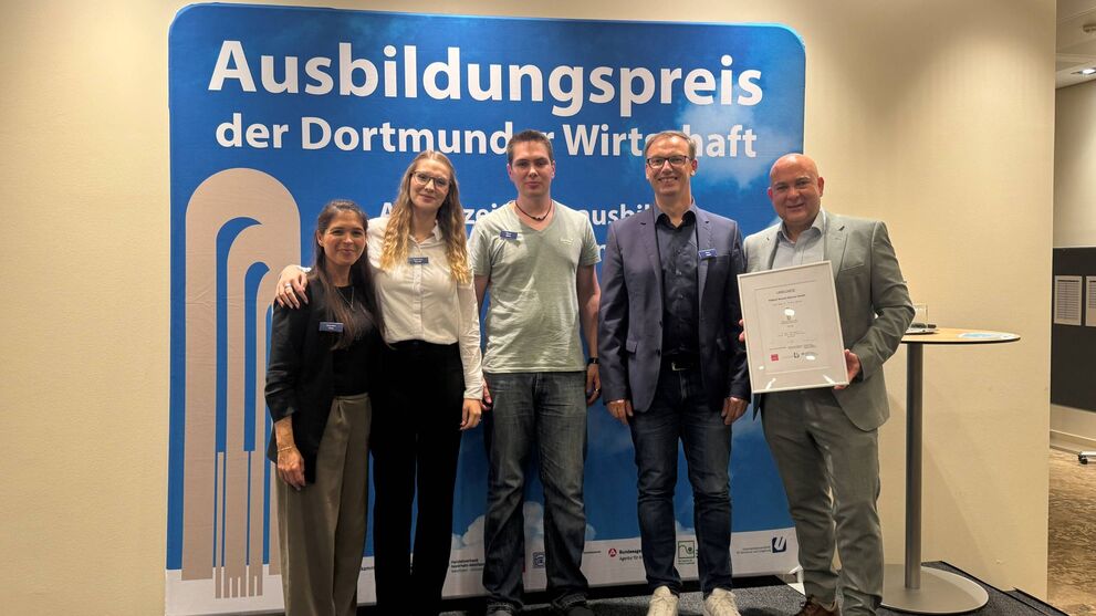 Gruppenfoto vor der Pressewand mit dem Ausbildungspreis. (v. l. n. r.): Gianina Pate, Anna Zoe Beda, Simon Gibbs, Heino Joppe und Kai Grefling.