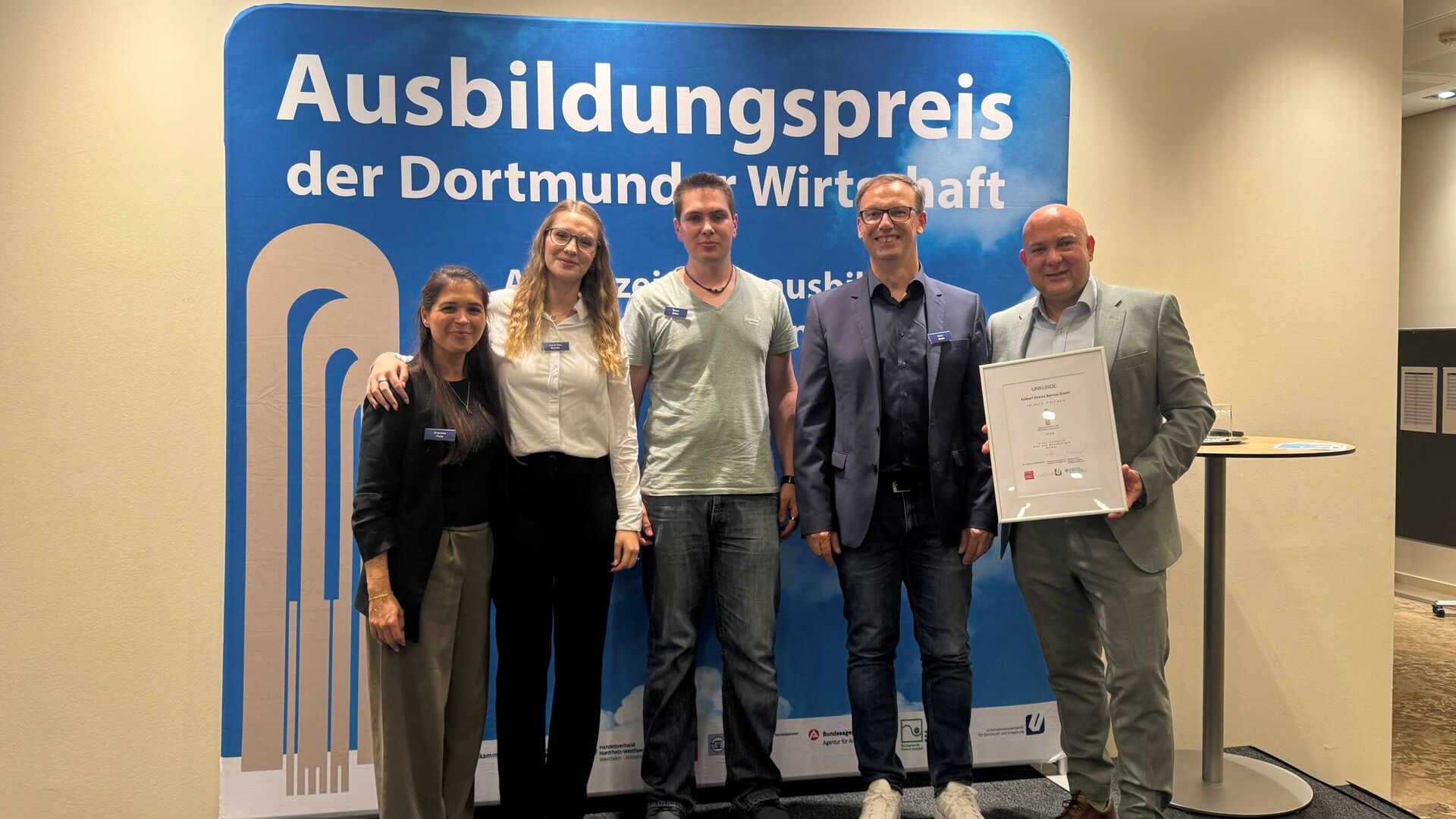 Gruppenfoto vor der Pressewand mit dem Ausbildungspreis. (v. l. n. r.): Gianina Pate, Anna Zoe Beda, Simon Gibbs, Heino Joppe und Kai Grefling.
