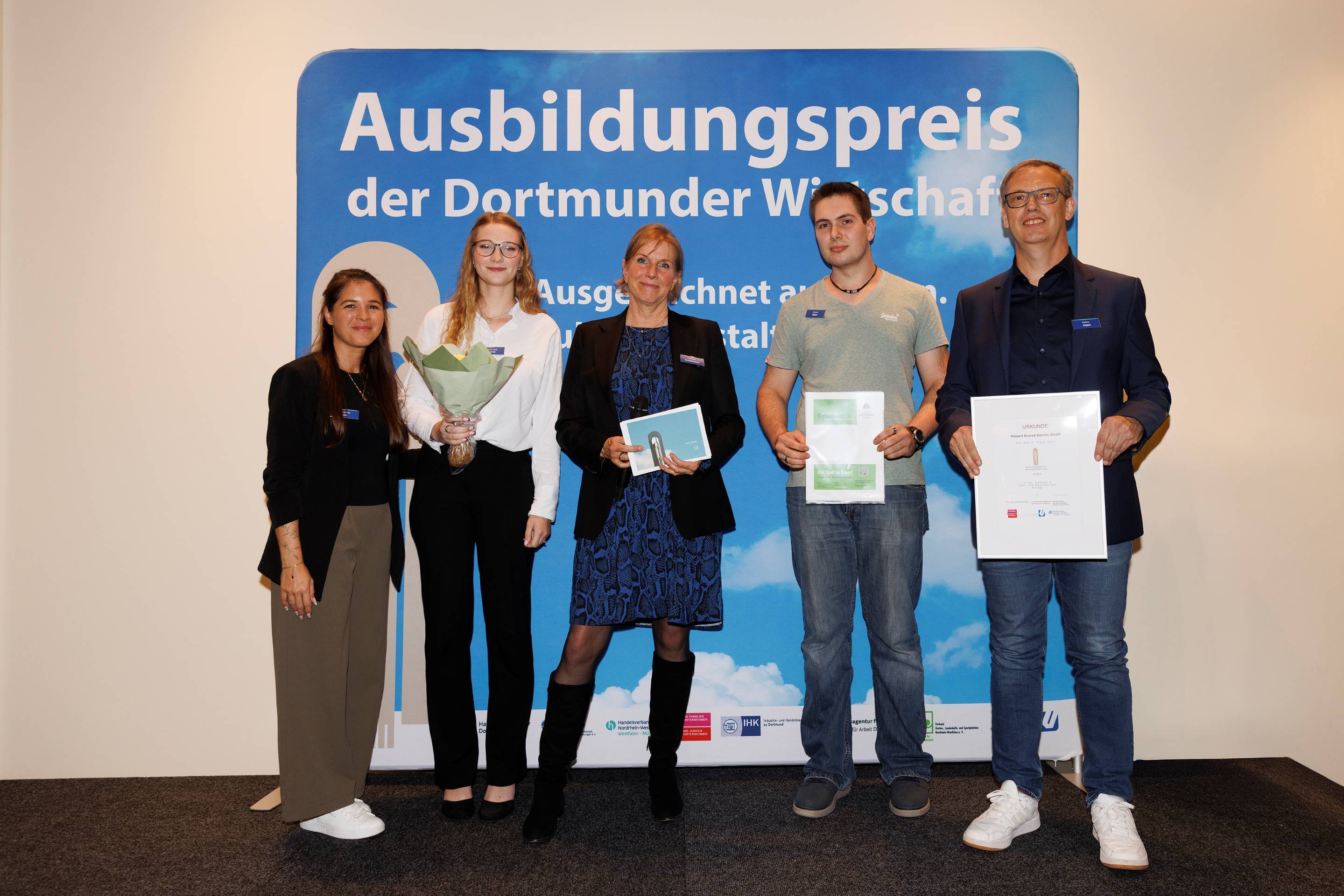 Gruppenfoto vor der Pressewand mit dem Ausbildungspreis. (v. l. n. r.): Gianina Pate, Anna Zoe Beda, Evelyn Knüffermann, Simon Gibbs und Heino Joppe.