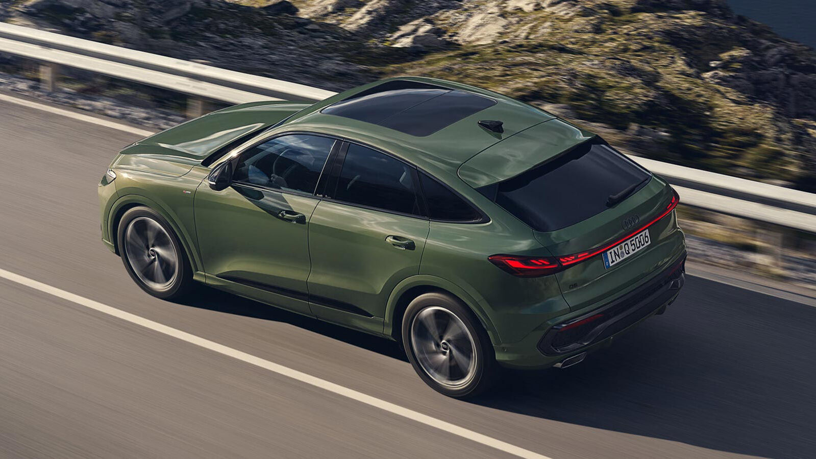 Audi Q5 Sportback in Distriktgrün Metallic fährt dynamisch eine Landstraße entlang, mit abfallender Coupé-Silhouette, Panorama-Glasdach, Led Heckleuchten und 20-Zoll-Felgen im 5-Speichen-Design.
