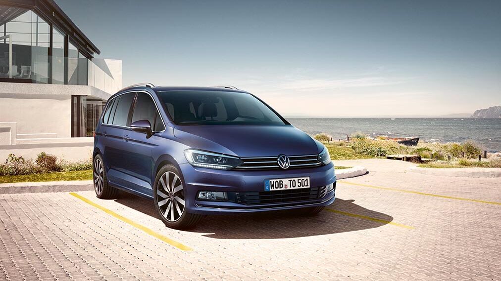 Der blaue VW Touran steht mit Blick auf das Meer vor moderner Architektur. Der flexible Innenraum macht ihn zum praktischen Begleiter für Familie und Alltag.